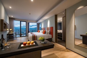 A modern hotel room with a large bed, a dining area with a view, and a balcony with mountains in the background.