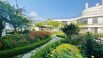 Un edificio moderno con fachada curva, rodeado de jardines bien cuidados con diversas plantas y flores.