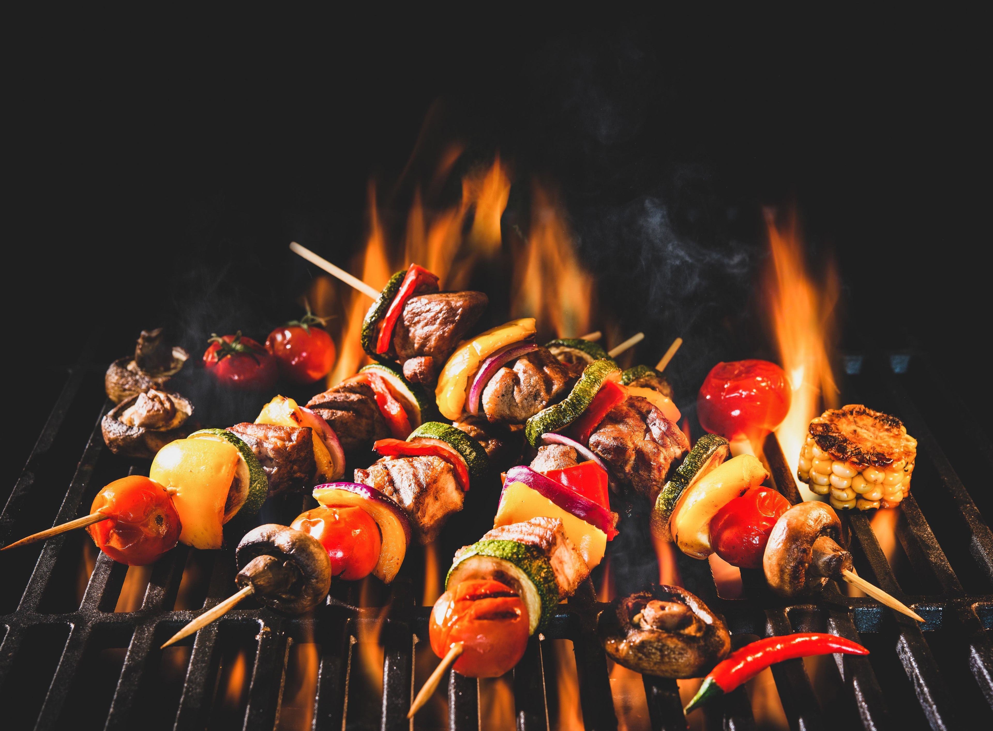 Diversas brochetas a la parrilla con verduras y carne, incluyendo tomates, pimientos y champiñones, sobre una barbacoa con llamas visibles.