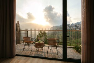 A balcony with wicker chairs and a table, overlooking a cityscape with mountains and a cloudy sky.