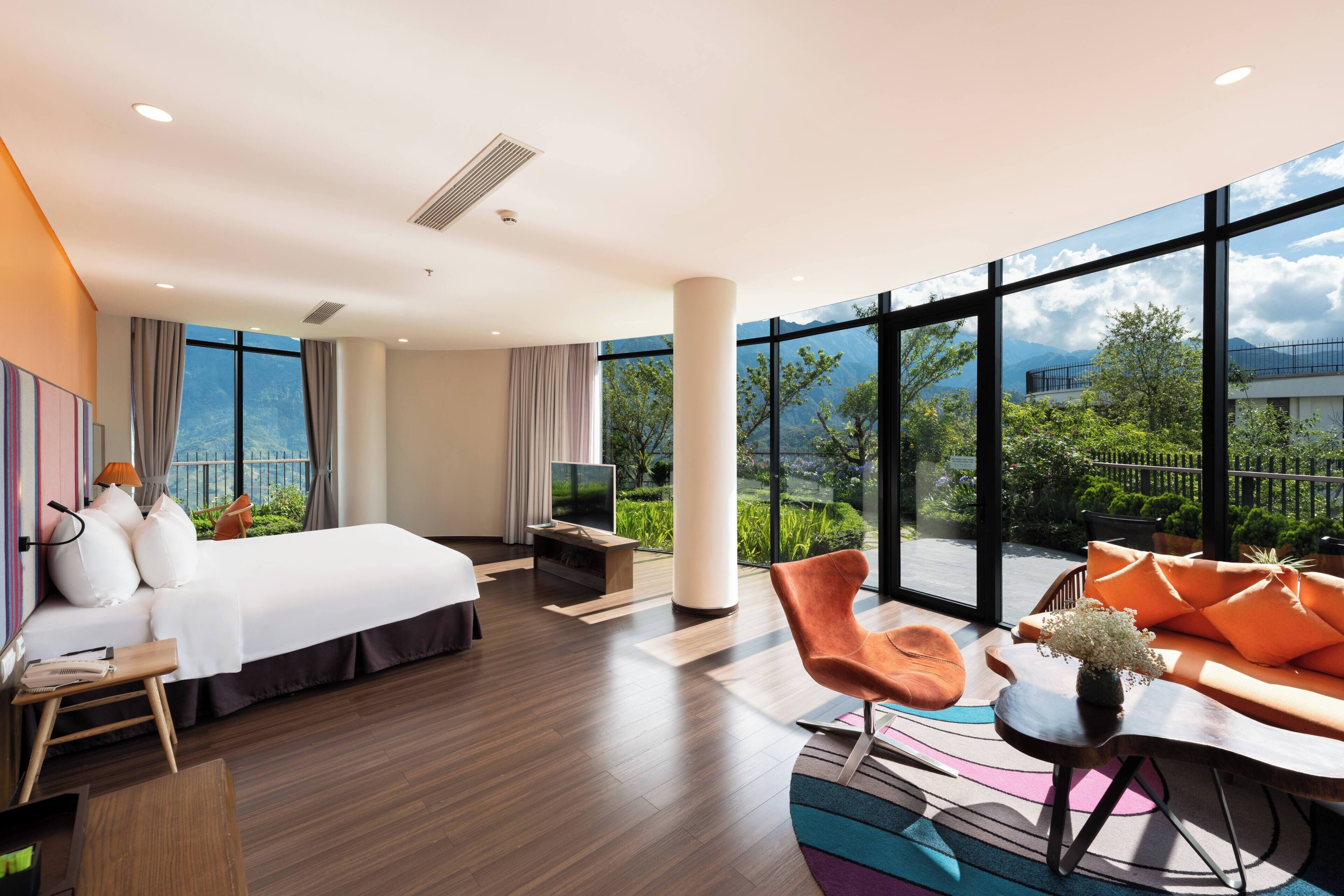 Habitación de hotel con una cama grande, televisor, sofá y vistas a zonas verdes y montañas.