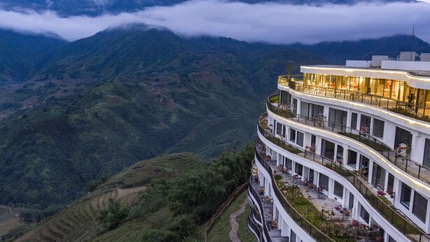 Hotel de vários andares com varanda curva, oferecendo vistas de uma paisagem montanhosa e nuvens.