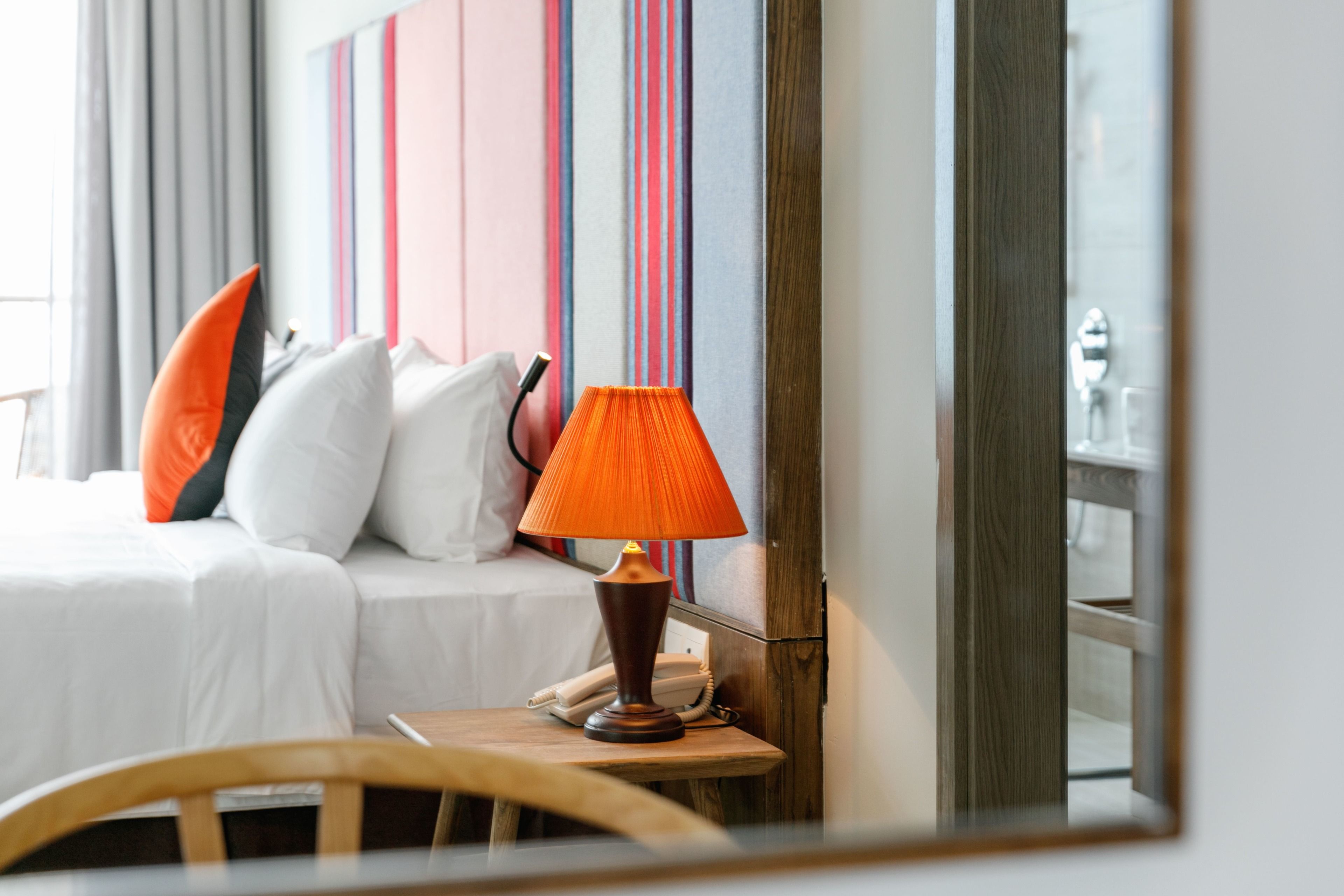 A hotel room with a bed, pillows, a lamp, a telephone, and a mirror reflecting part of the room.