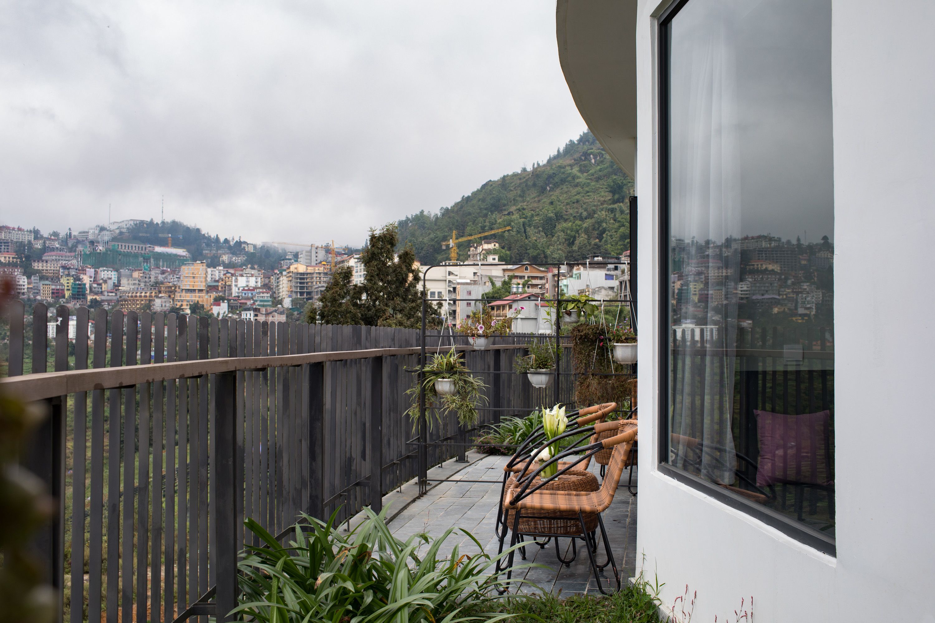 Un balcón con sillas de mimbre, plantas en macetas y vistas de un paisaje urbano con edificios y colinas al fondo.