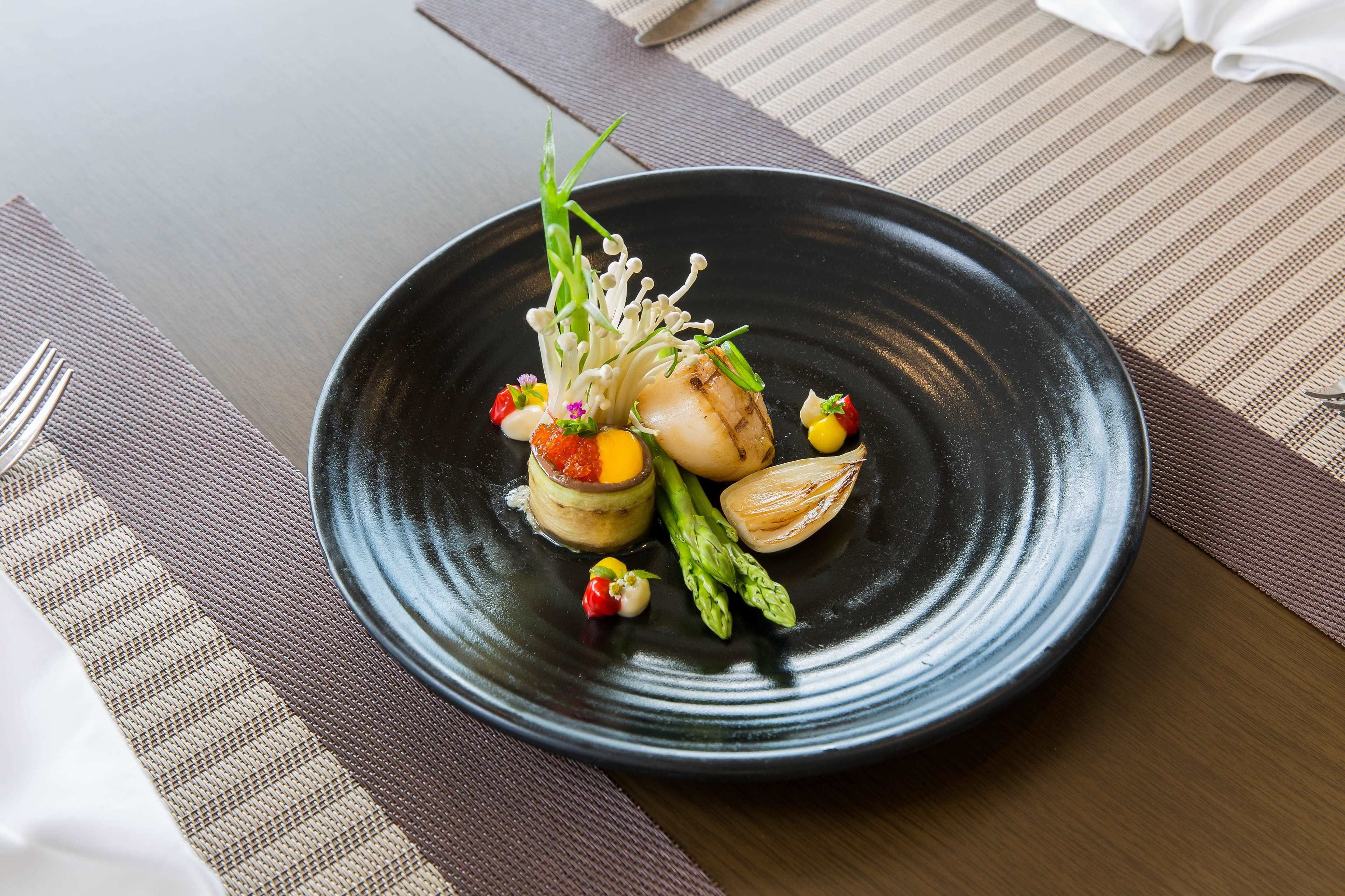 A black plate with a scallop, asparagus, and small vegetables on a table with a striped placemat.