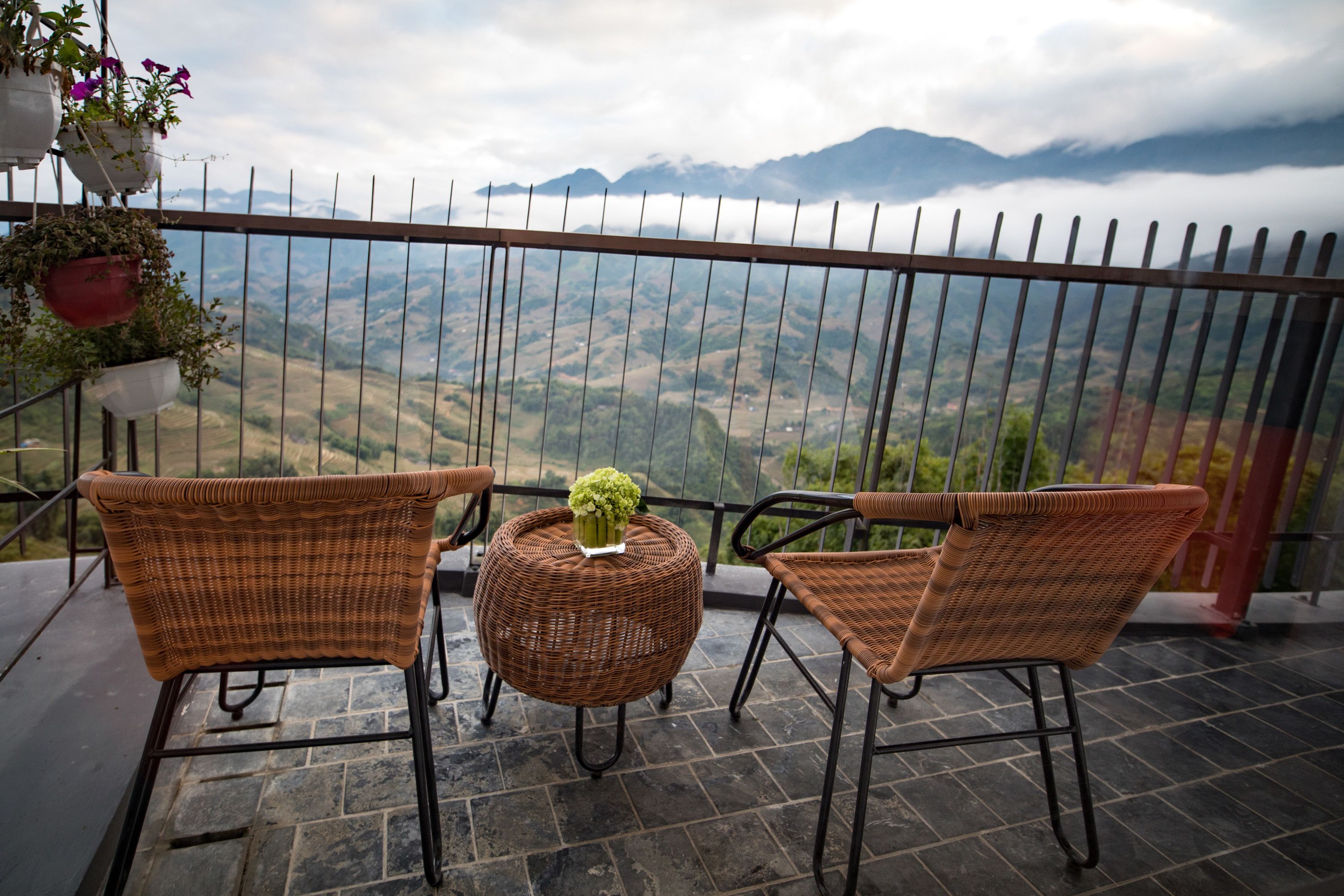 Een balkon met rieten stoelen en een tafel, met uitzicht op een bergachtig landschap.