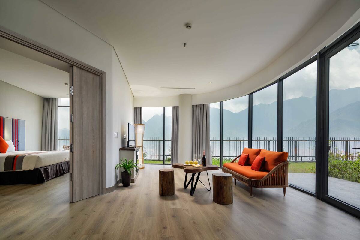 Ein modernes Hotelzimmer mit großem Balkon, einem Bett, einer Couch, einem Esstisch und Blick auf Berge.