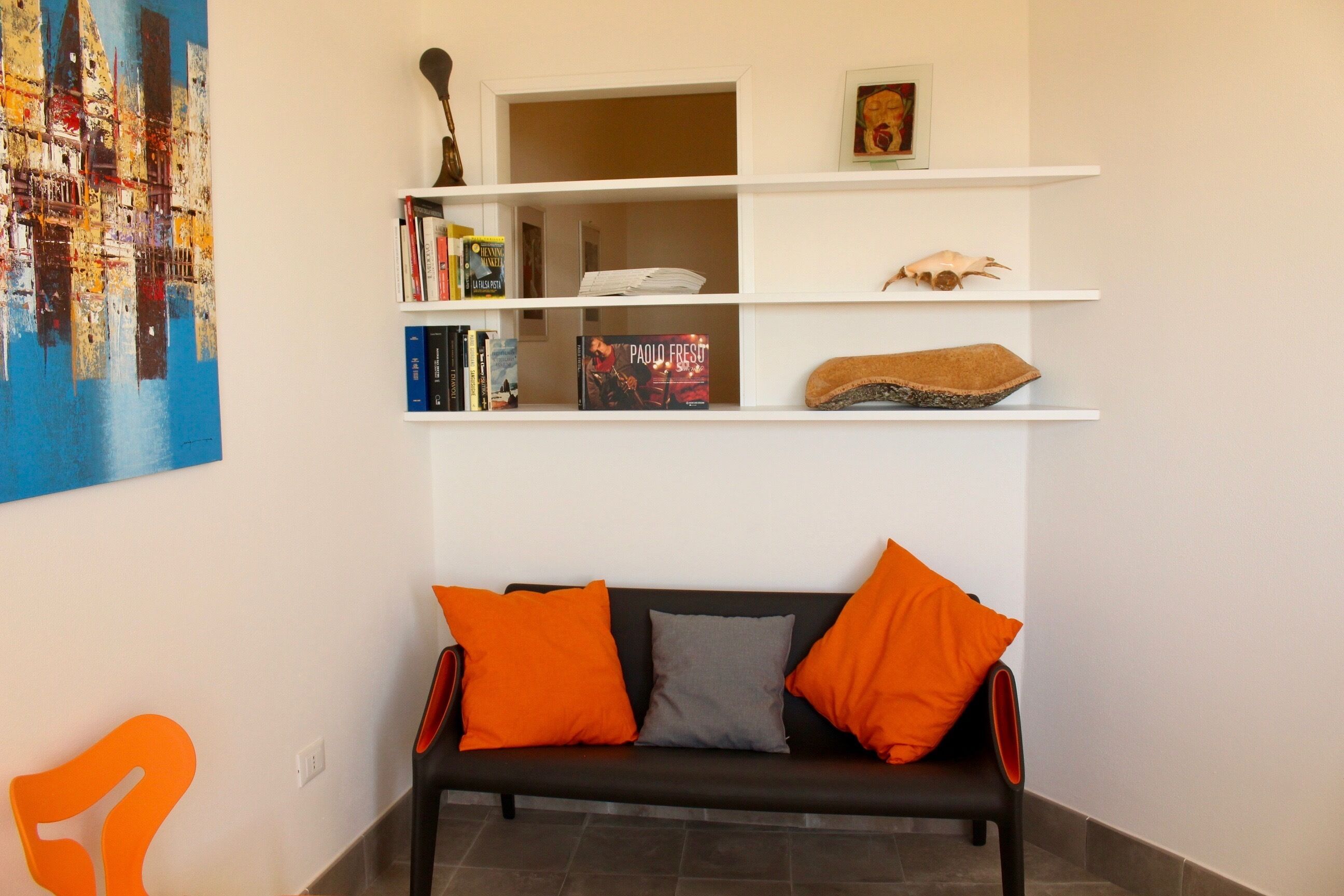 A living room with a black sofa, orange and gray cushions, a bookshelf with books and decorative items, and a cityscape painting on the wall.