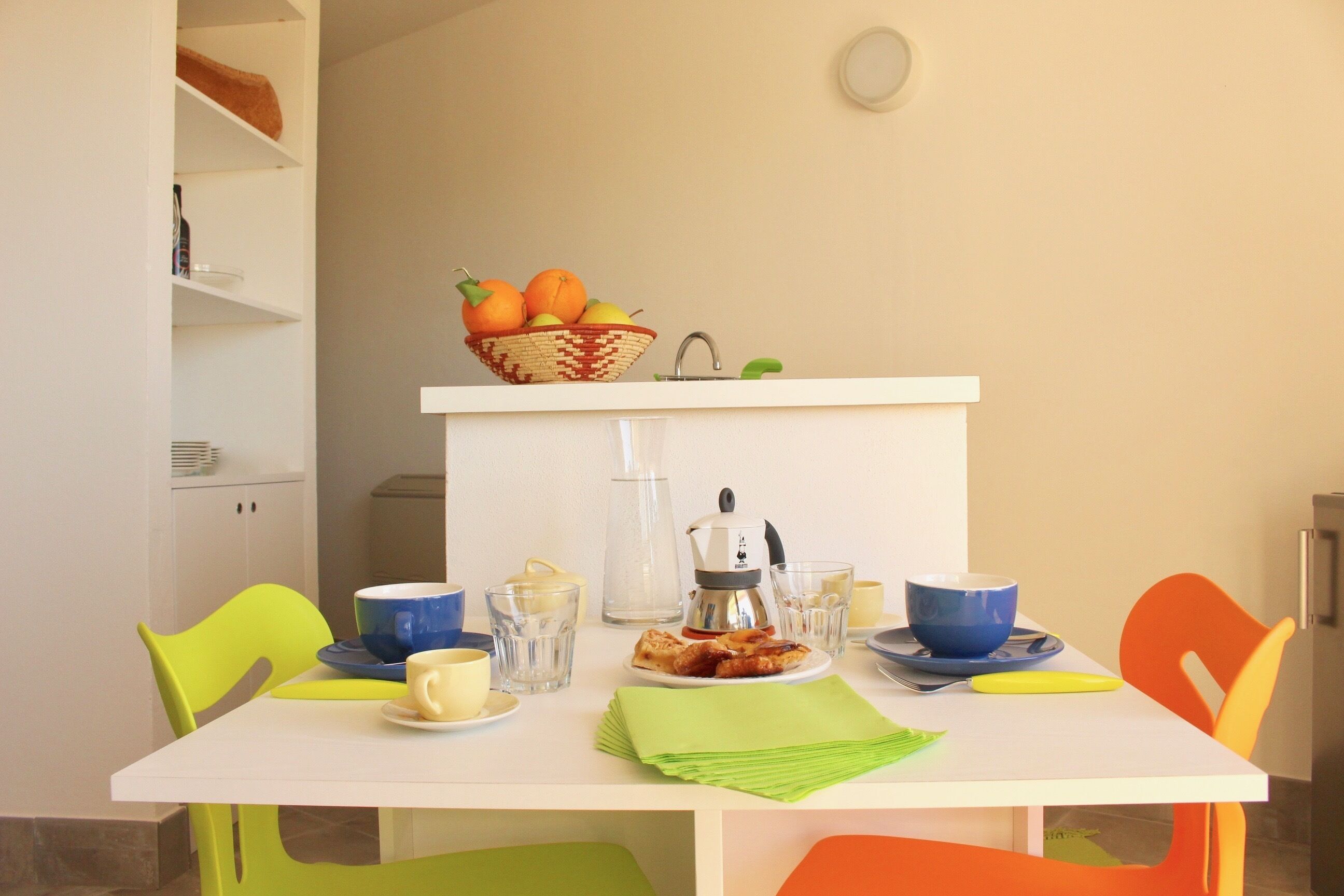 Un coin repas avec une table blanche, des chaises colorées, un panier de fruits, une machine à café, ainsi que différentes tasses et assiettes.