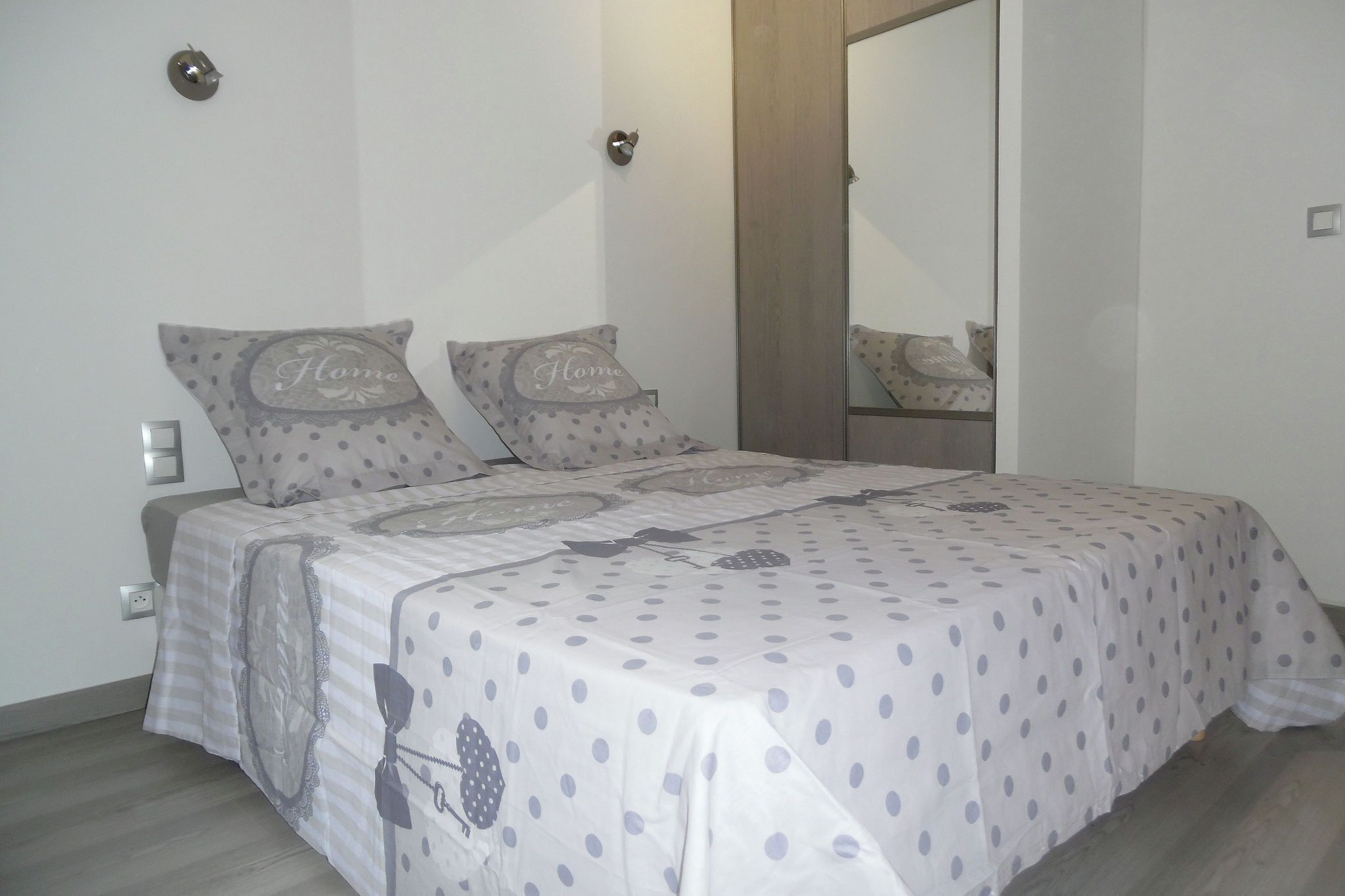 A bedroom with a bed covered in a polka dot bedspread, two pillows with the word "Home" on them, a wooden door, and a mirror on the wall.