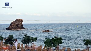 Un área de comedor frente a la playa con mesas y sillas, con vistas al mar y una formación rocosa al fondo.