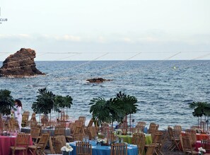 Un área de comedor frente a la playa con mesas y sillas, con vistas al mar y una formación rocosa al fondo.