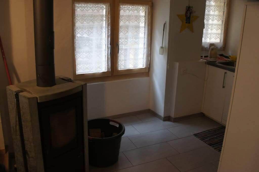 A small kitchen with a wood-burning stove, a window with lace curtains, and a star decoration on the wall.