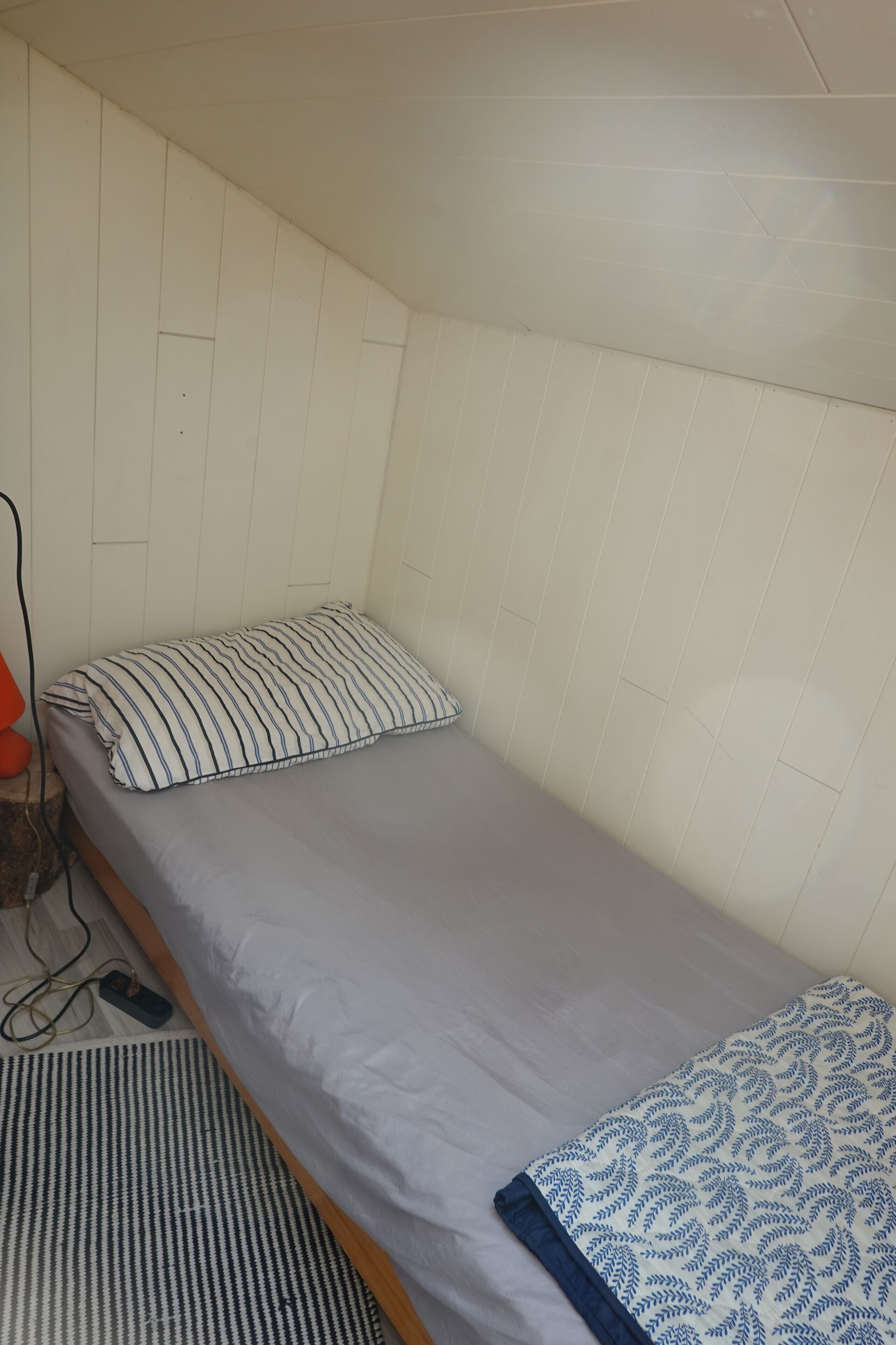 Ein kleines Zimmer mit einem Einzelbett, einer schlichten weißen Wand und einem Bett mit einem gestreiften Kopfkissen.