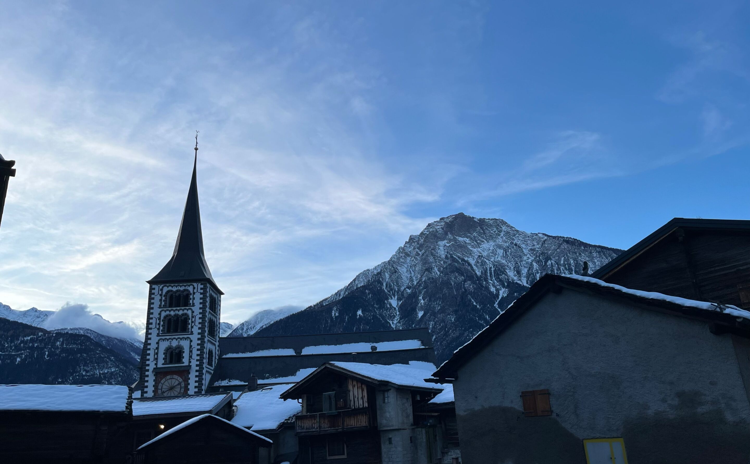Un village de montagne enneigé, avec une église dotée d’un clocher élancé et d’une tour horloge.