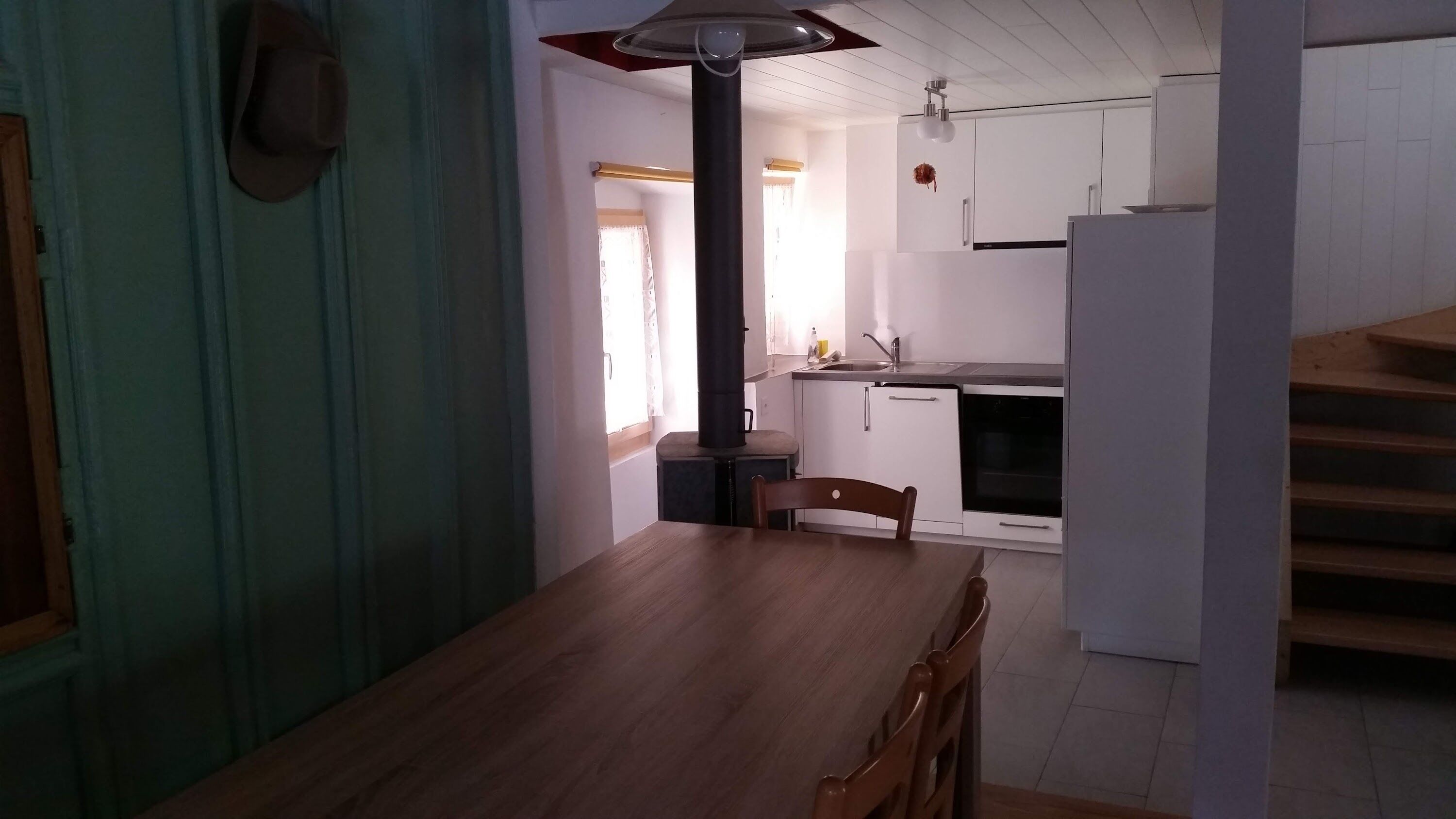 Ruokailutila, jossa on puinen pöytä ja tuolit, valkoisilla kaapeilla ja liedellä varustettu keittiö sekä toiselle tasolle johtavat portaat.
