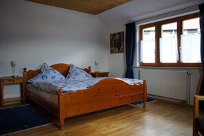 Ein Schlafzimmer mit einem hölzernen Bett, einem Nachttisch, einer Lampe, einem Fenster mit Vorhängen und einer Heizung.