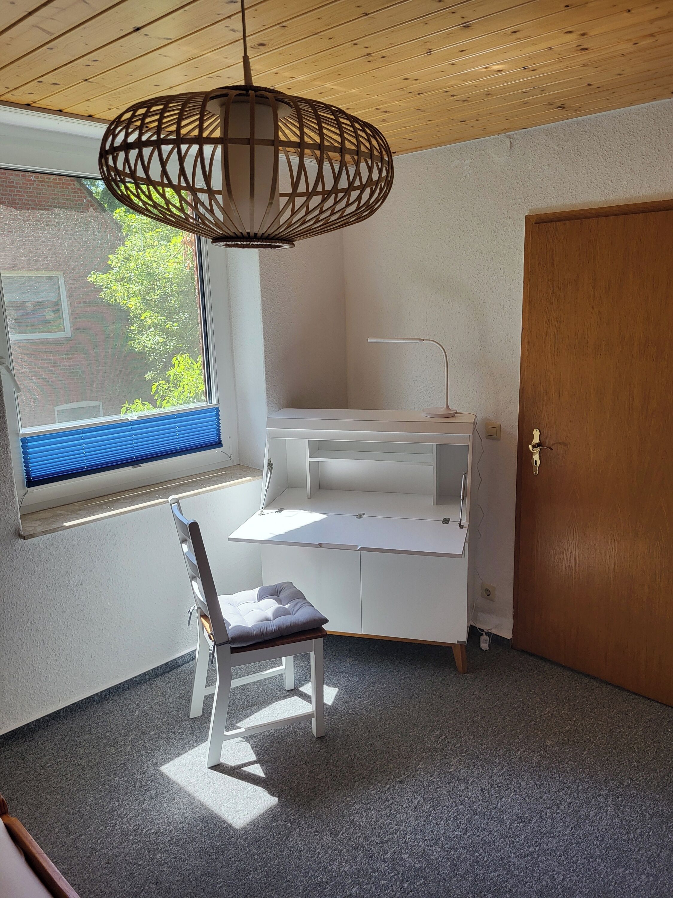 Een kamer met een houten plafond, een wit bureau met lades, een stoel, een raam met jaloezieën en een deur.