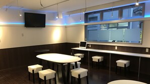 A modern conference room with a large oval table, smaller round tables, and white cushioned stools. A flat-screen TV is mounted on the wall, and there is a window with blinds. - HOTEL OWL Tokyo (Tokyo)