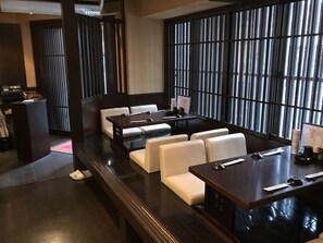 A modern restaurant with dark wood furniture, white cushioned seating, and a large window with vertical blinds. - HOTEL OWL Tokyo (Tokyo)