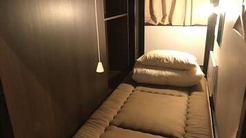 A small, single-bed room with a simple design, featuring a bed with a beige mattress and pillows, a hanging light fixture, and a small wardrobe.