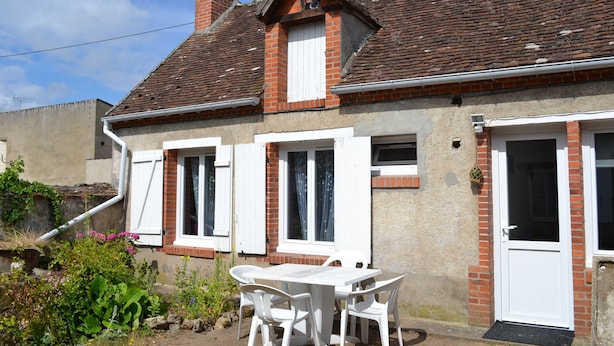 Une maison avec une façade en briques, des volets blancs et un toit en tuiles. On peut voir une table et des chaises blanches à l’extérieur.