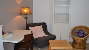 Une pièce comprenant une table blanche, un fauteuil sombre agrémenté d’un coussin rose, une chaise en osier avec un coussin violet, une haute armoire blanche et un lampadaire doté d’un abat-jour à motifs.
