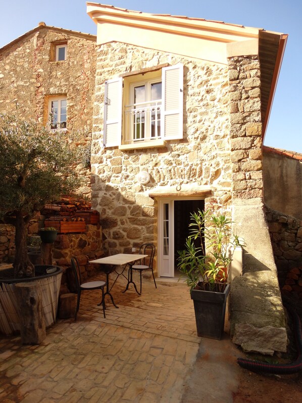 Un bâtiment en pierre avec une porte en bois, une fenêtre avec des volets et un petit balcon agrémenté d’une table et de chaises.