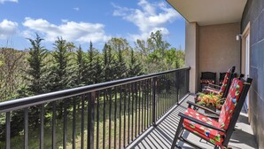 Un balcon doté d’une balustrade en métal, deux chaises recouvertes d’un tissu à motifs floraux, et une vue sur les arbres et le ciel.