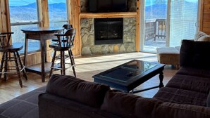 A cozy cabin interior with a stone fireplace, wooden walls, a large window, and a balcony. - KastleRock Brand new 5 min to Slopes PoolTable Jacuzzi VIEWS PRIVATE sleep15 (Banner Elk)