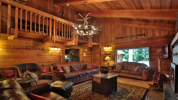 L’intérieur chaleureux d’un chalet en rondins, avec un escalier en bois, des canapés en cuir, un lustre et une grande fenêtre donnant sur le paysage extérieur.