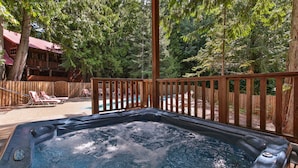 Un bain à remous doté d’une terrasse en bois et d’une clôture en bois, entouré d’arbres et bordé par un bâtiment au toit rouge.
