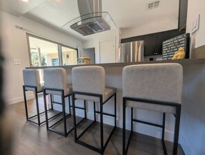 A modern kitchen with a stainless steel refrigerator, a built-in oven, and a range hood. There are three bar stools with black frames and light-colored seats. A decorative item is placed on the counter. - CENTRALLY LOCATED WITH HOT TUB: Zen Garden - Your Sedona Oasis Awaits (Sedona)