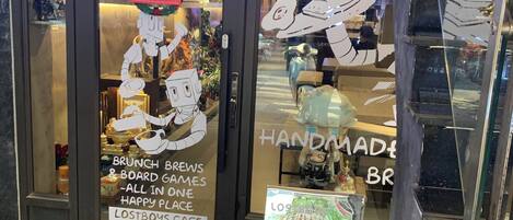 Exterior del Lostboys Cafe, con un felpudo que dice 'WELCOME' y un cartel anunciando brunch y juegos de mesa.