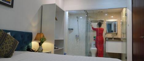 A hotel room with a bed, a bedside table with a lamp, a vase with flowers, a shower area with a person, and a bathroom with a mirror.