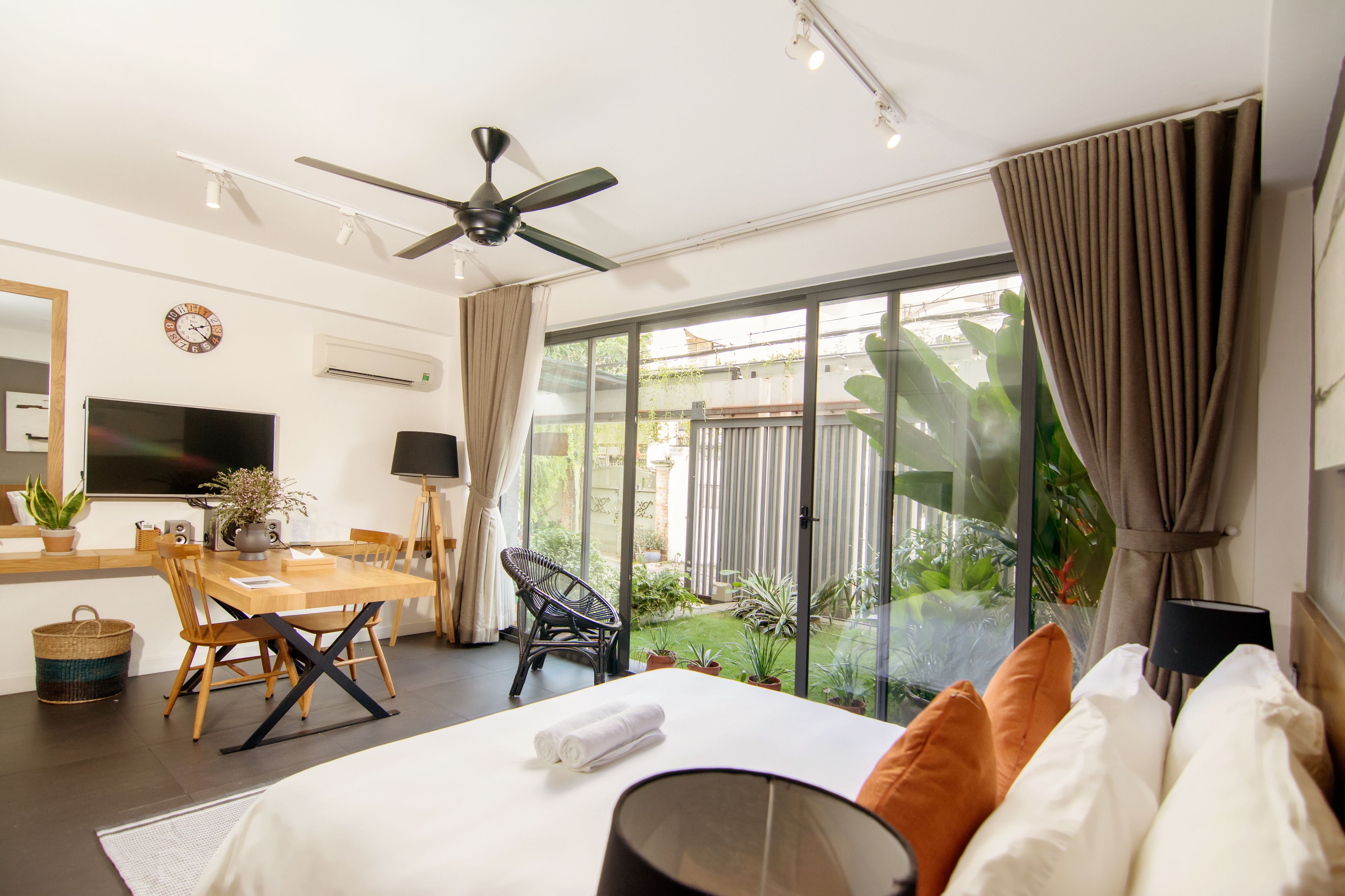 A hotel room with a bed, a dining table, chairs, a ceiling fan, a TV, and a view of the garden.