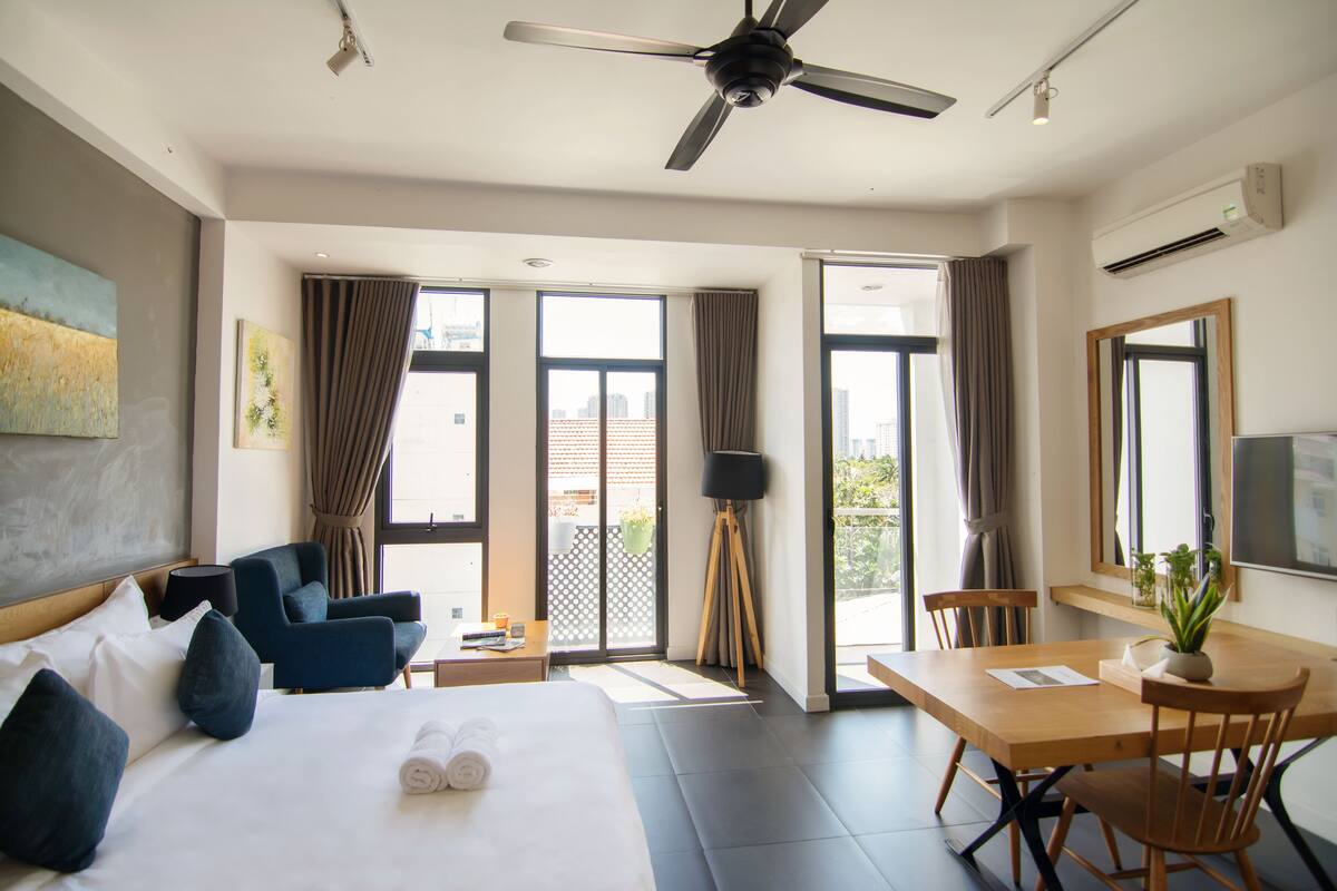 A hotel room with a bed, a desk, a chair, a ceiling fan, and large windows.