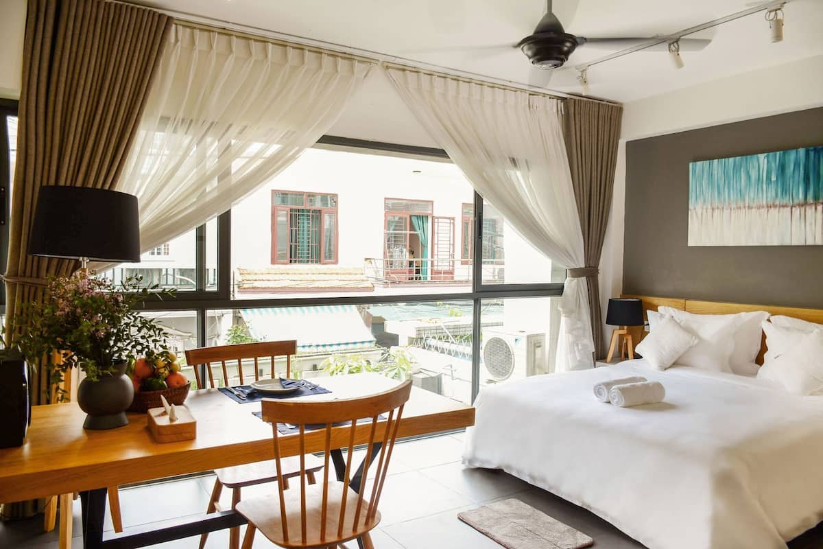 A hotel room with a bed, a dining table with chairs, a potted plant, and a view of buildings outside.