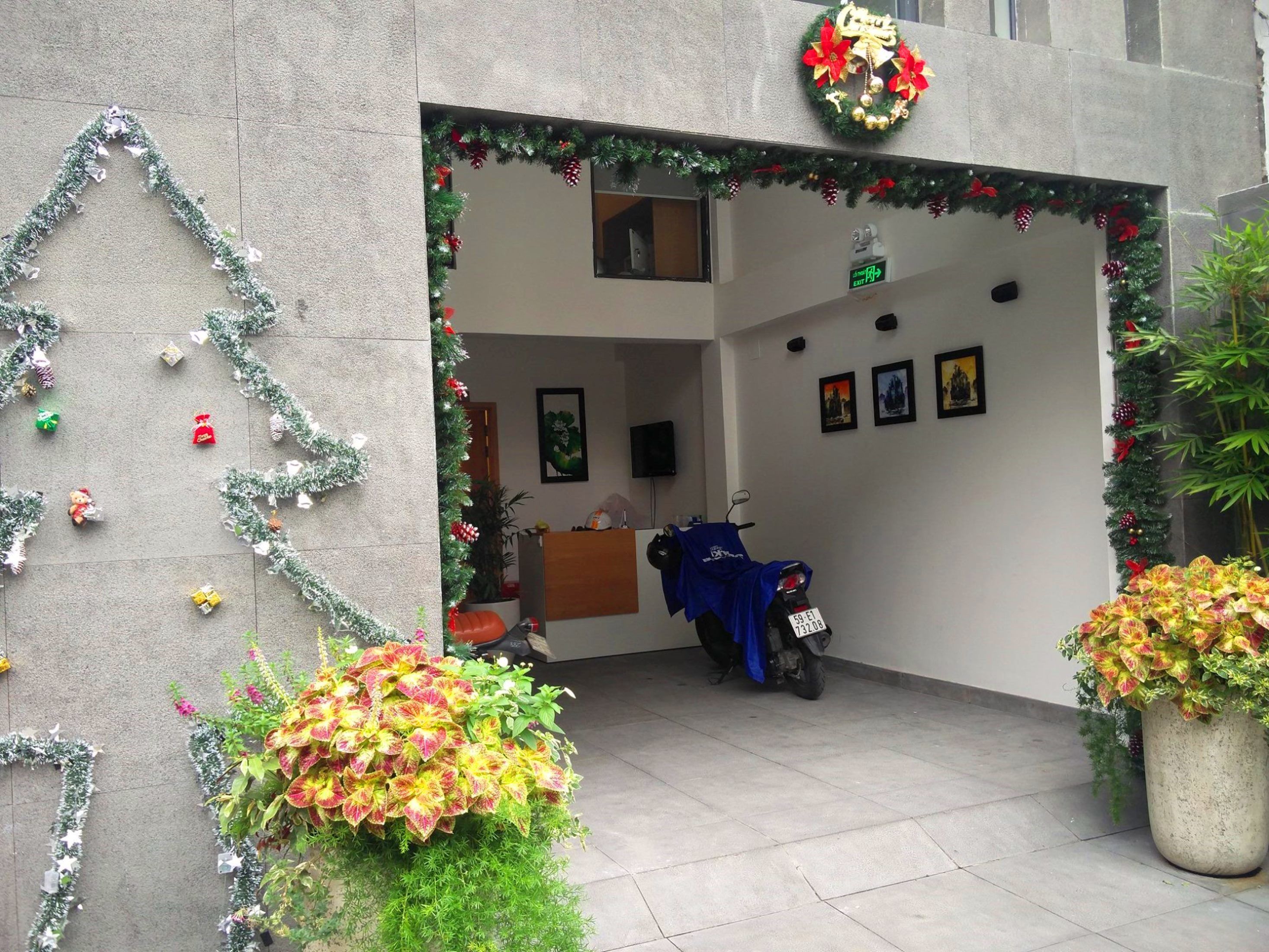 Fachada de hotel decorada con guirnaldas navideñas y un adorno circular, que muestra un mostrador de recepción, una motocicleta y plantas de interior.