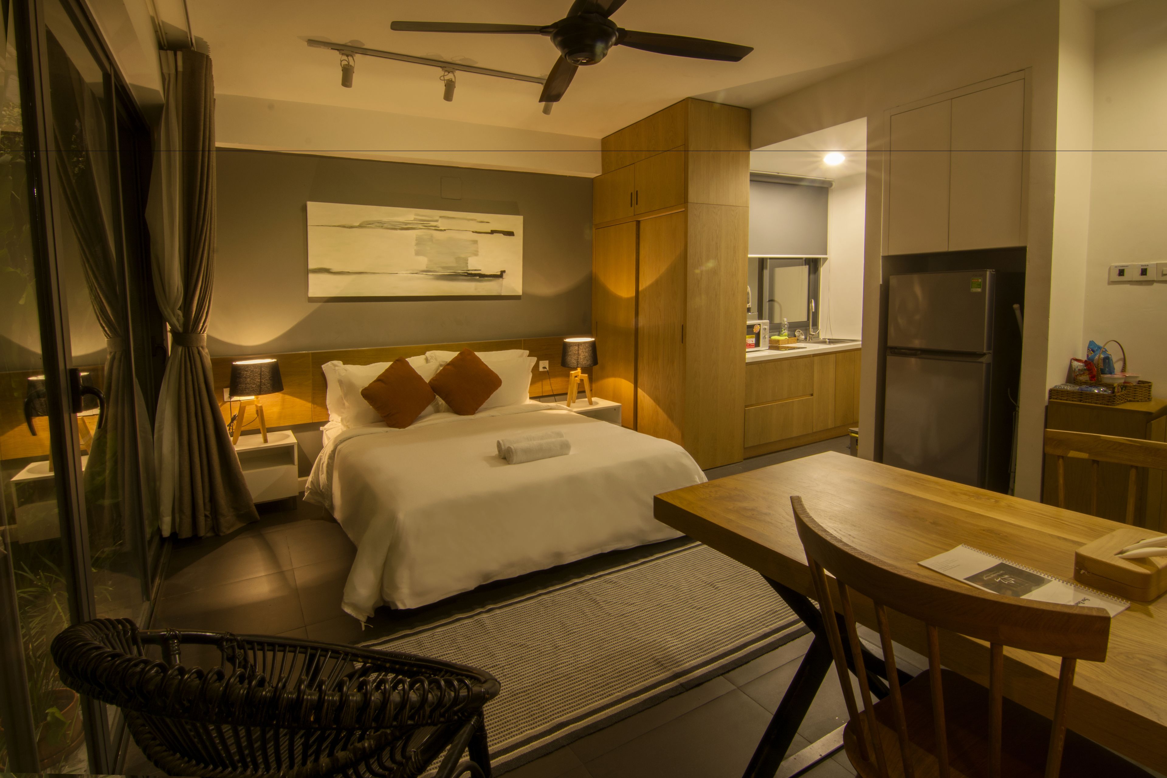 A hotel room with a bed, bedside tables, a desk, a chair, a ceiling fan, a refrigerator, and a window with curtains.