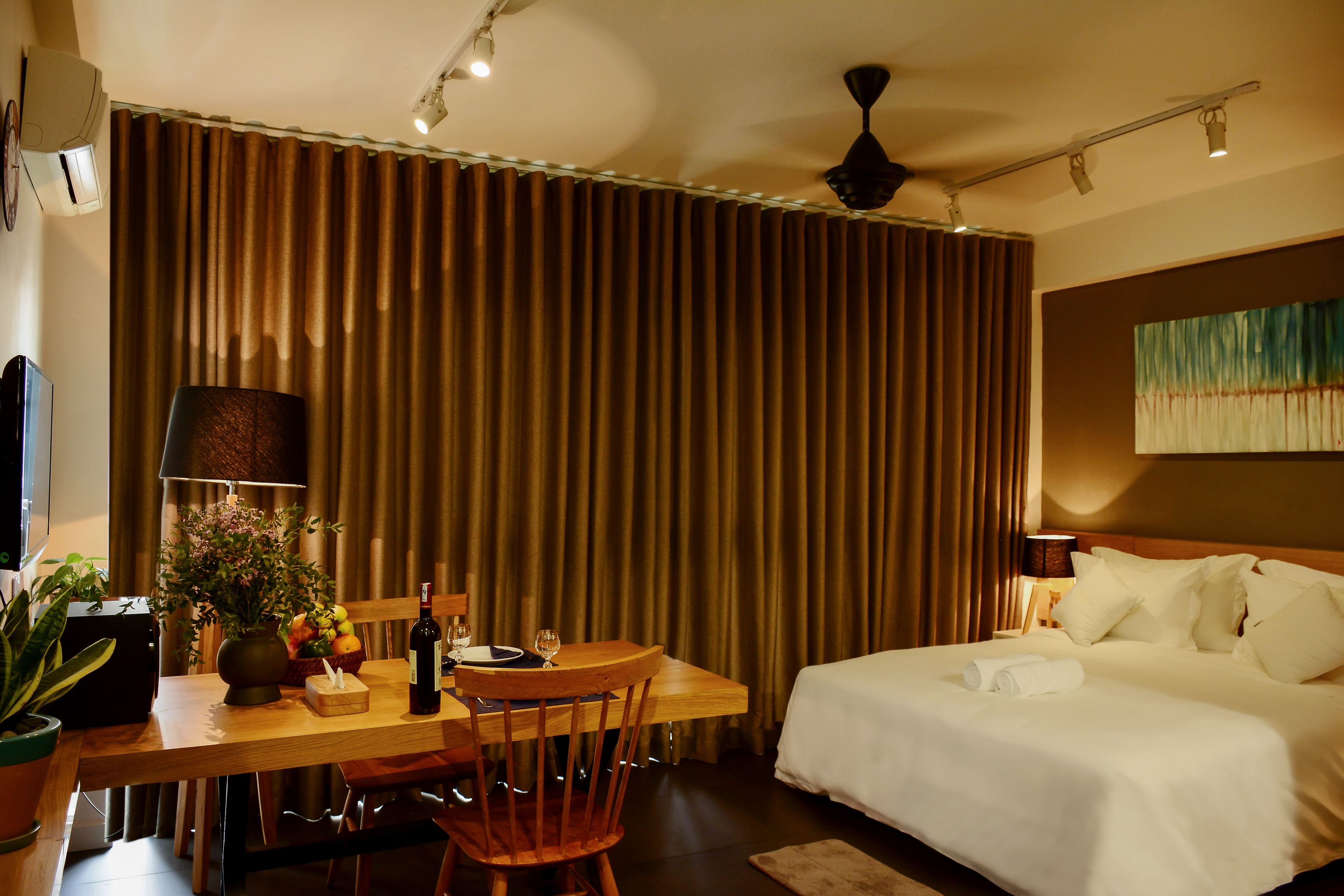 A hotel room with a bed, a desk with a chair, a lamp, a vase with flowers, and a large window with curtains.