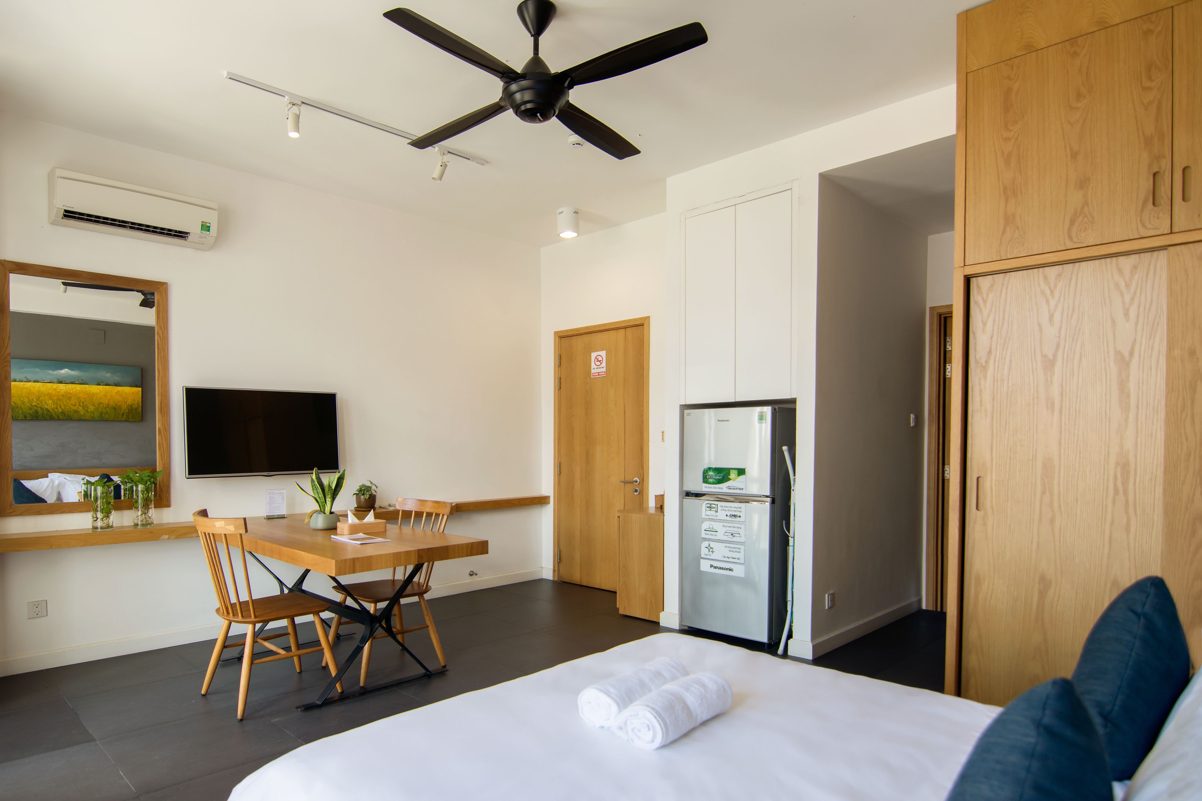 Ett hotellrum med en säng, ett skrivbord med stolar, ett kylskåp, en takfläkt och en stor spegel.
