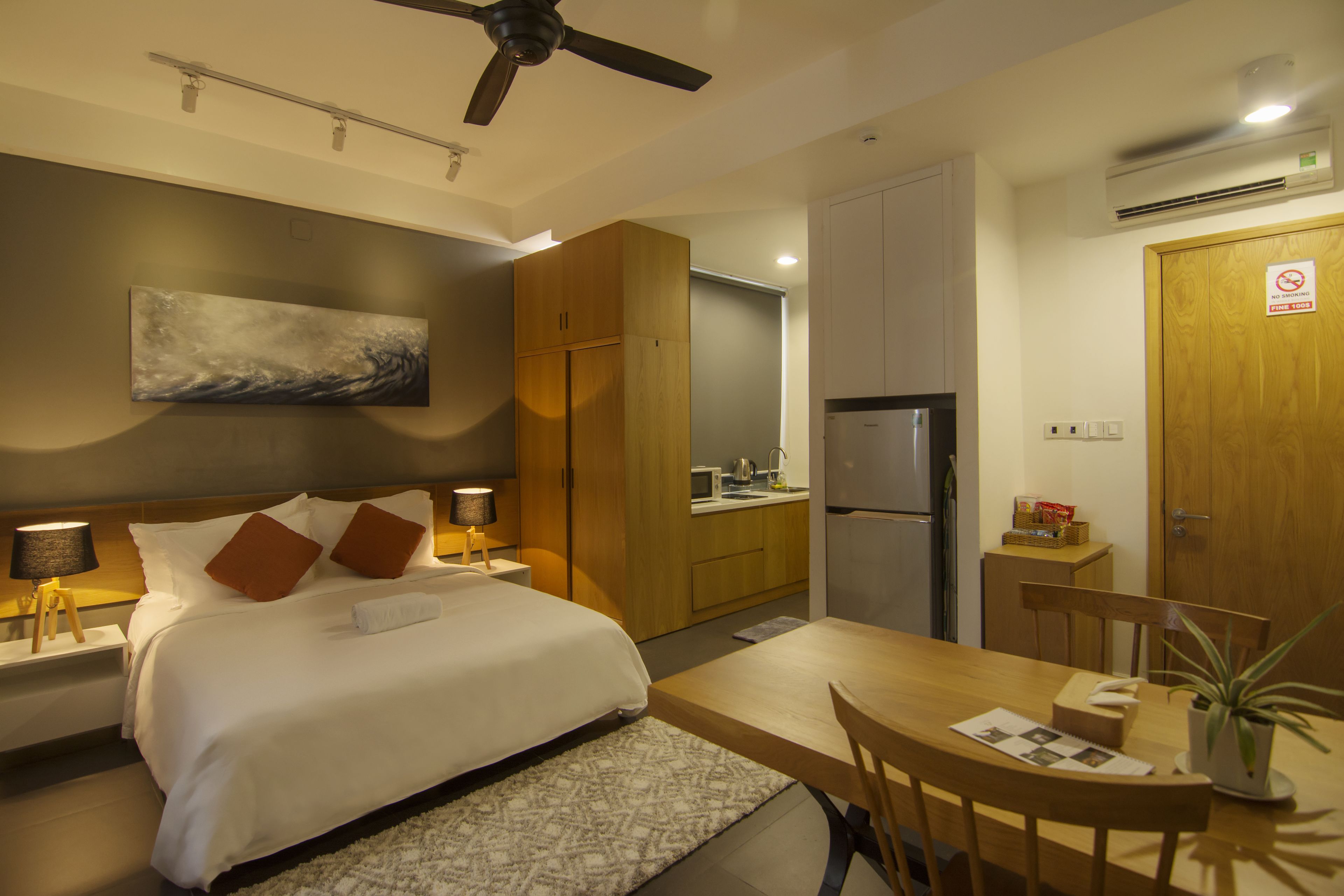 Una camera d'albergo moderna con un letto, lampade da comodino, una ventola a soffitto, un quadro, un angolo cottura e un tavolo da pranzo.