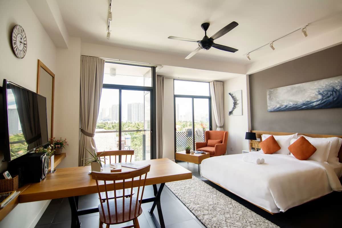 A hotel room with a bed, a desk with a chair, a television, a ceiling fan, and large windows.