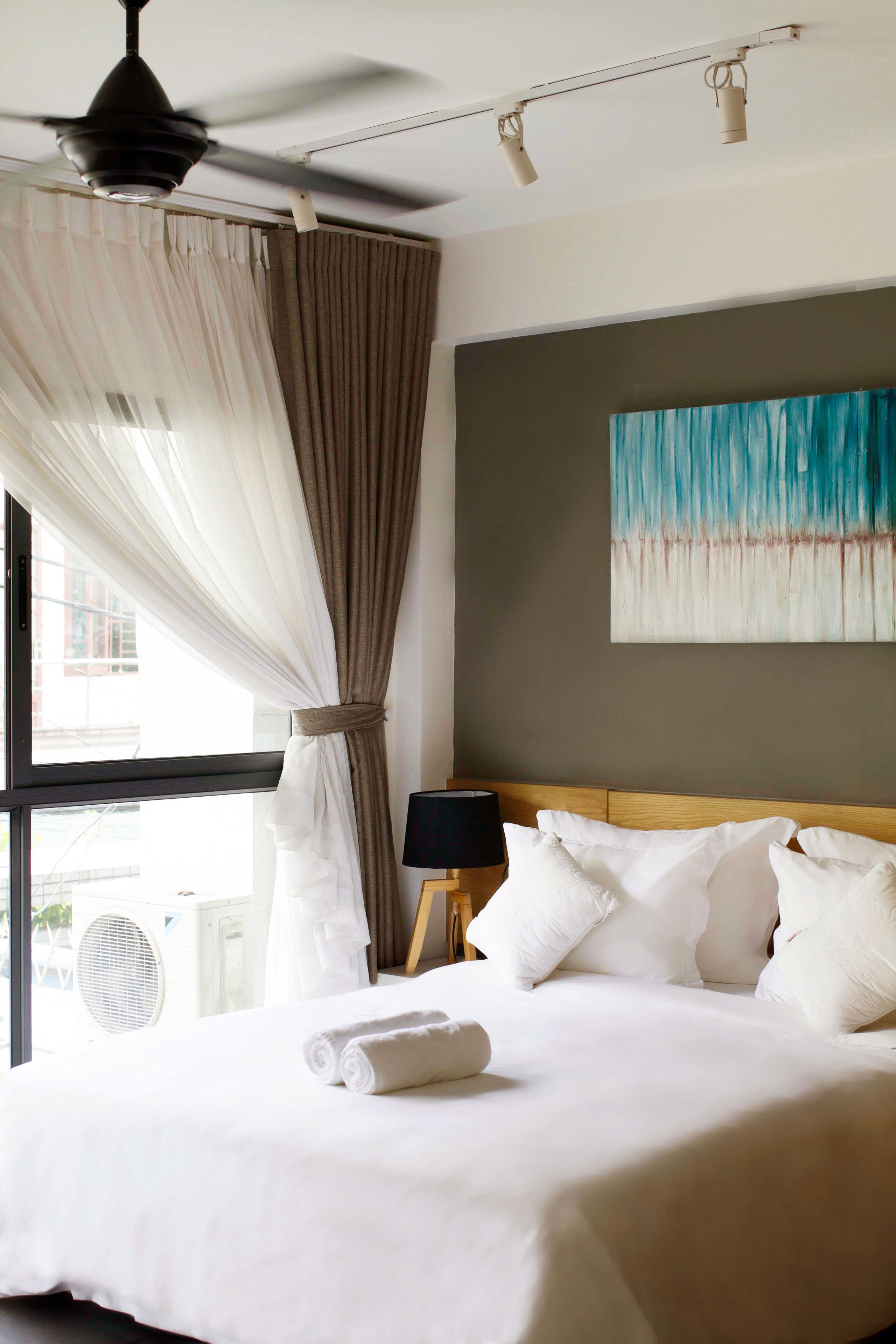 Ein Hotelzimmer mit einem Bett, weißen Vorhängen, einem Deckenventilator, einem Gemälde an der Wand und einem Fenster mit Klimaanlage.