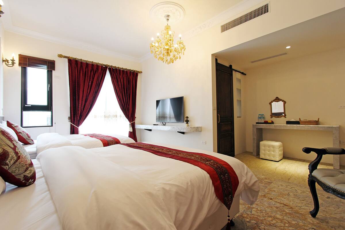 A hotel room with two beds, a TV, a desk, a chair, and a chandelier.
