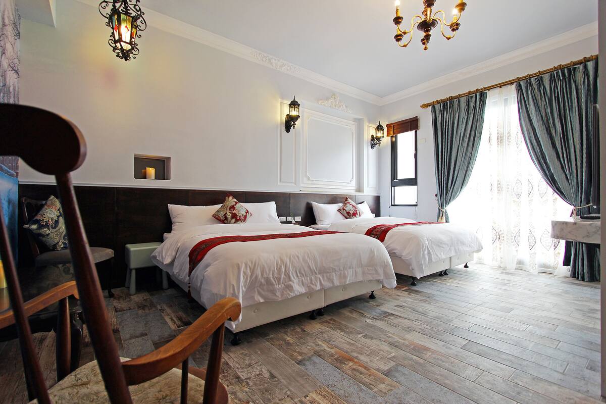 A hotel room with two beds, wooden flooring, a chandelier, and a large window with curtains.