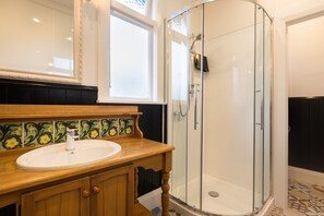Un baño con tocador de madera, lavamanos, cabina de ducha con mampara de vidrio y salpicadero de azulejos estampados.