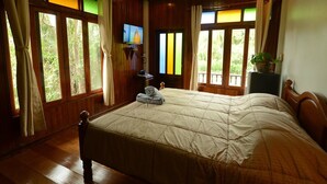 A bedroom with a large bed, wooden walls, a television, and a view of greenery through the windows. - River Jam Amphawa (Amphawa)