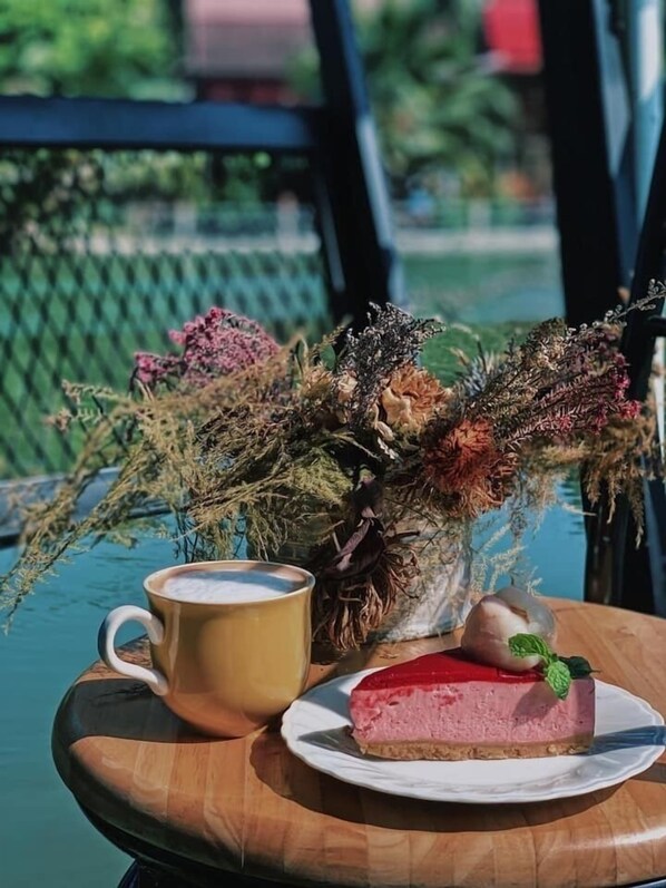 A table with a cup of coffee, a plate with a slice of cheesecake, and a vase with dried flowers.